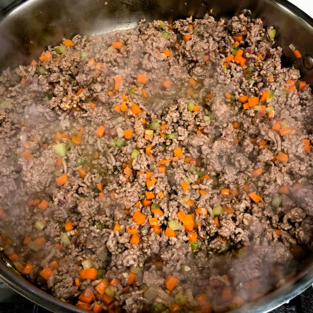 Bolognese sauce ingredients cooking the ground beef, carrots, onion, and celery in a large skillet.