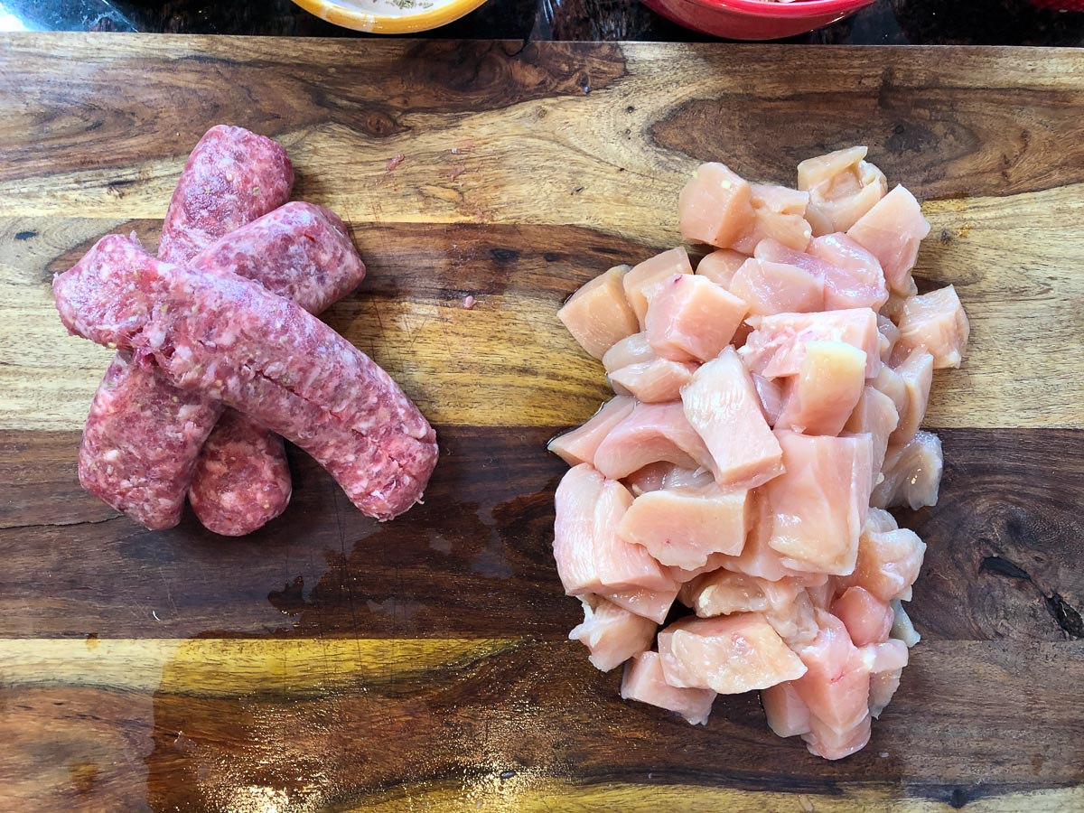 raw chicken cut into cubes next to raw Italian sausage on a cutting board